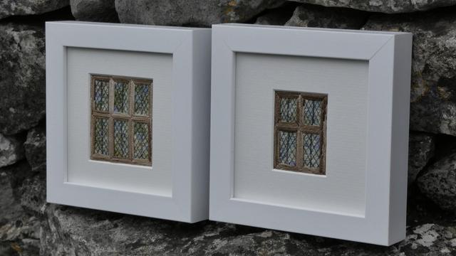 Two small, stitched artworks depicting mullioned, leaded windows. Framed in white and resting against a stone wall