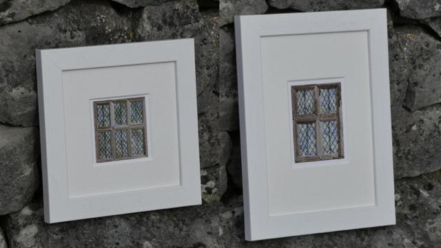 Two small prints, taken from stitched artworks depicting mullioned, leaded windows. Framed in white and resting against a stone wall