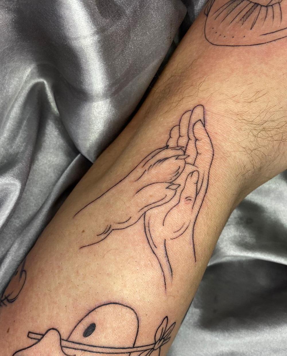 Foto do meu braço direito em um fundo prateado mostrando uma tatuagem preta de uma pata de cachorro to ando uma mão humana, como se estivesse se cumprimentando.