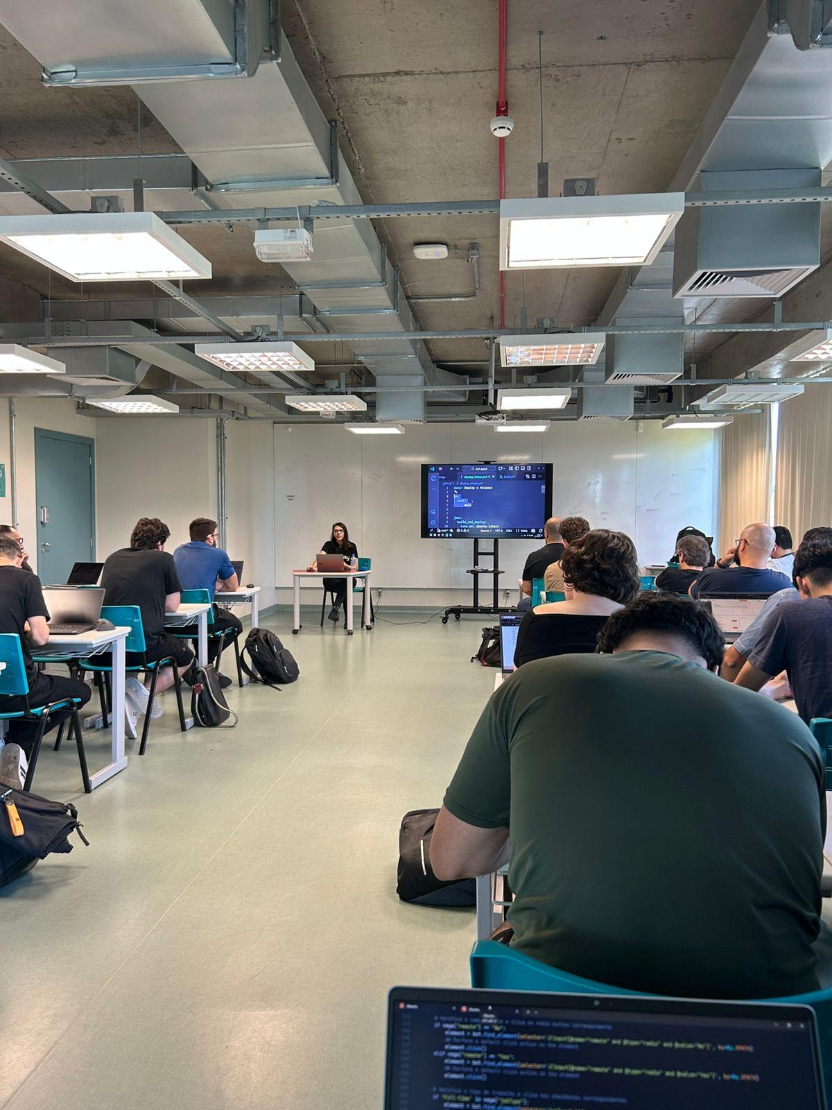 Foto de sala de aula. Foto tirada do fundo da sala, varias pessoas sentadas, eu na frente da turma, ensinando um tutorial sobre automação. Do meu lado uma tela grande mostrando códigos.