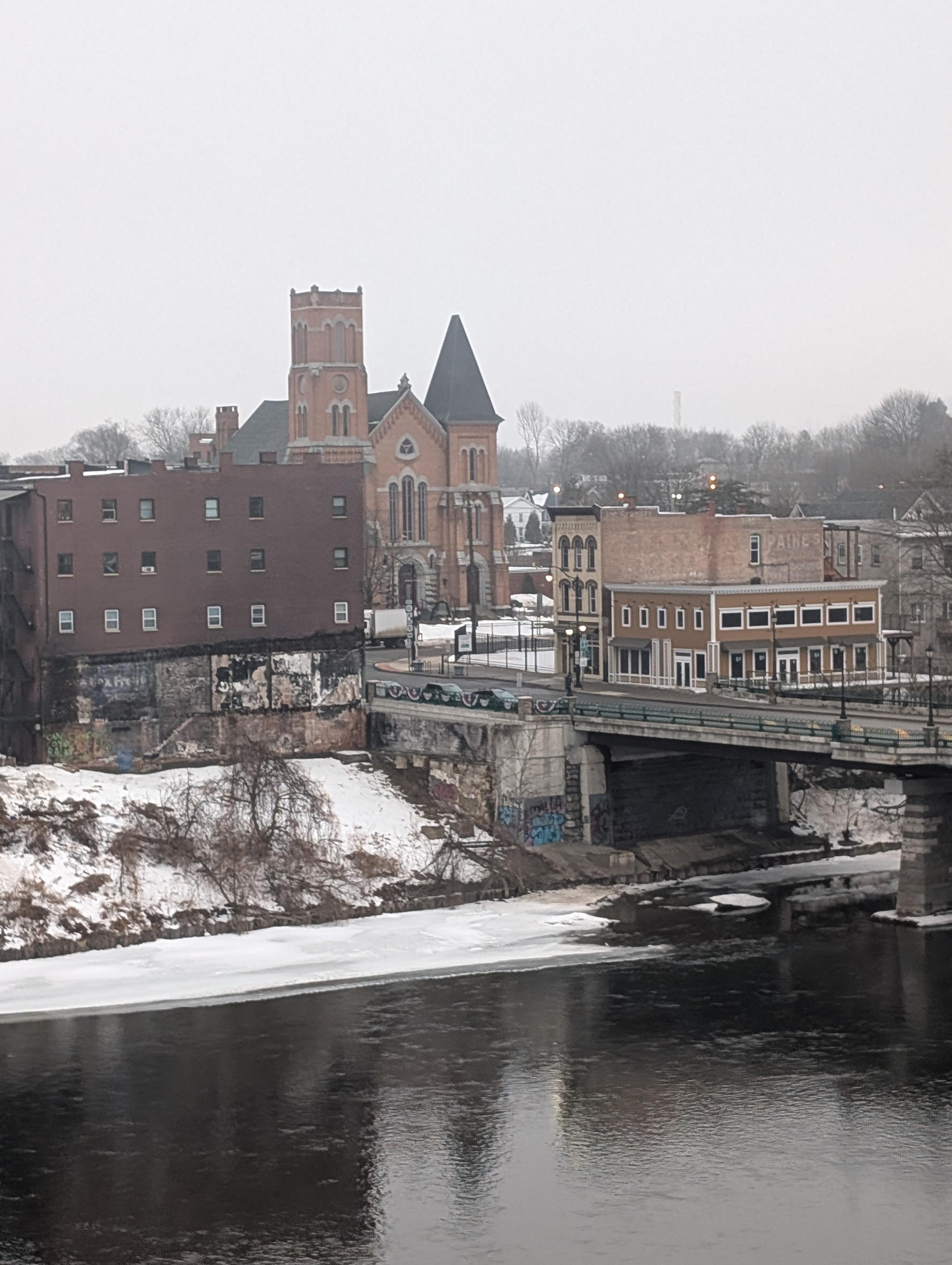 Uma paisagem urbana em tons frios capturada durante o inverno. Em primeiro plano, as águas escuras de um rio refletem os edifícios, com placas de gelo e neve acumulada nas margens. Uma ponte de concreto atravessa o rio, conectando a área a uma rua com edifícios de tijolos de diversos estilos. No centro, destaca-se uma imponente igreja em estilo neogótico, construída em tijolos avermelhados, com uma torre quadrada e um telhado pontiagudo escuro. O cenário é completado por árvores sem folhas e um céu cinzento e uniforme, transmitindo uma atmosfera calma e melancólica.