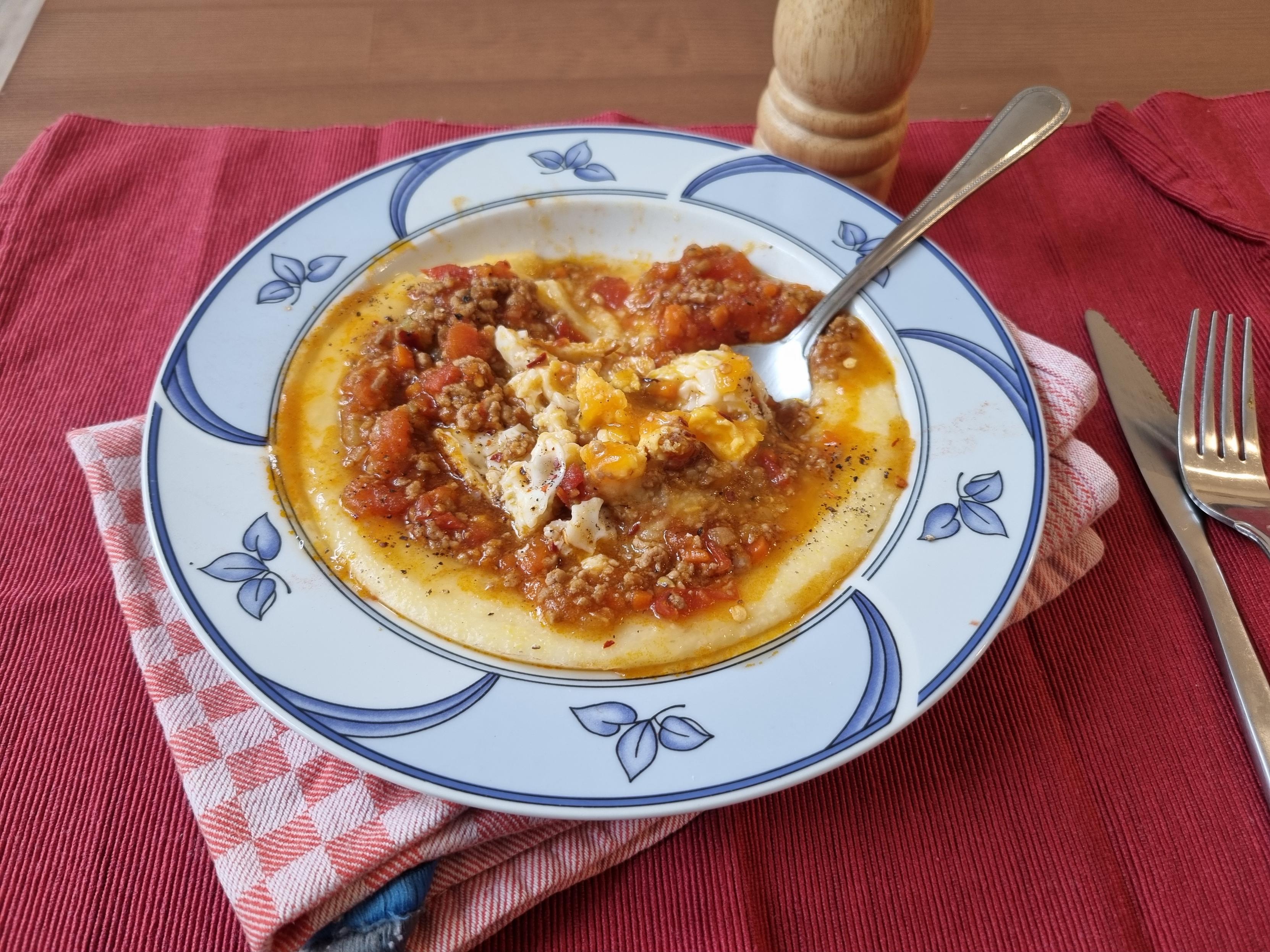 Prato fundo branco com desenhos azuis nas bordas, os desenhos são 6 ramos com 3 folhas cada ao redor do prato. O prato contém polenta coberta com molho bolonhesa e um ovo frito picado. Eles está em cima de um prato de pano xadrez branco e vermelho que por sua vez está em cima de um pano vermelho escuro.
Dentro do prato está uma colher, e ao lado direito um garfo e uma faca.
Ao fundo um moedor de pimenta de madeira.