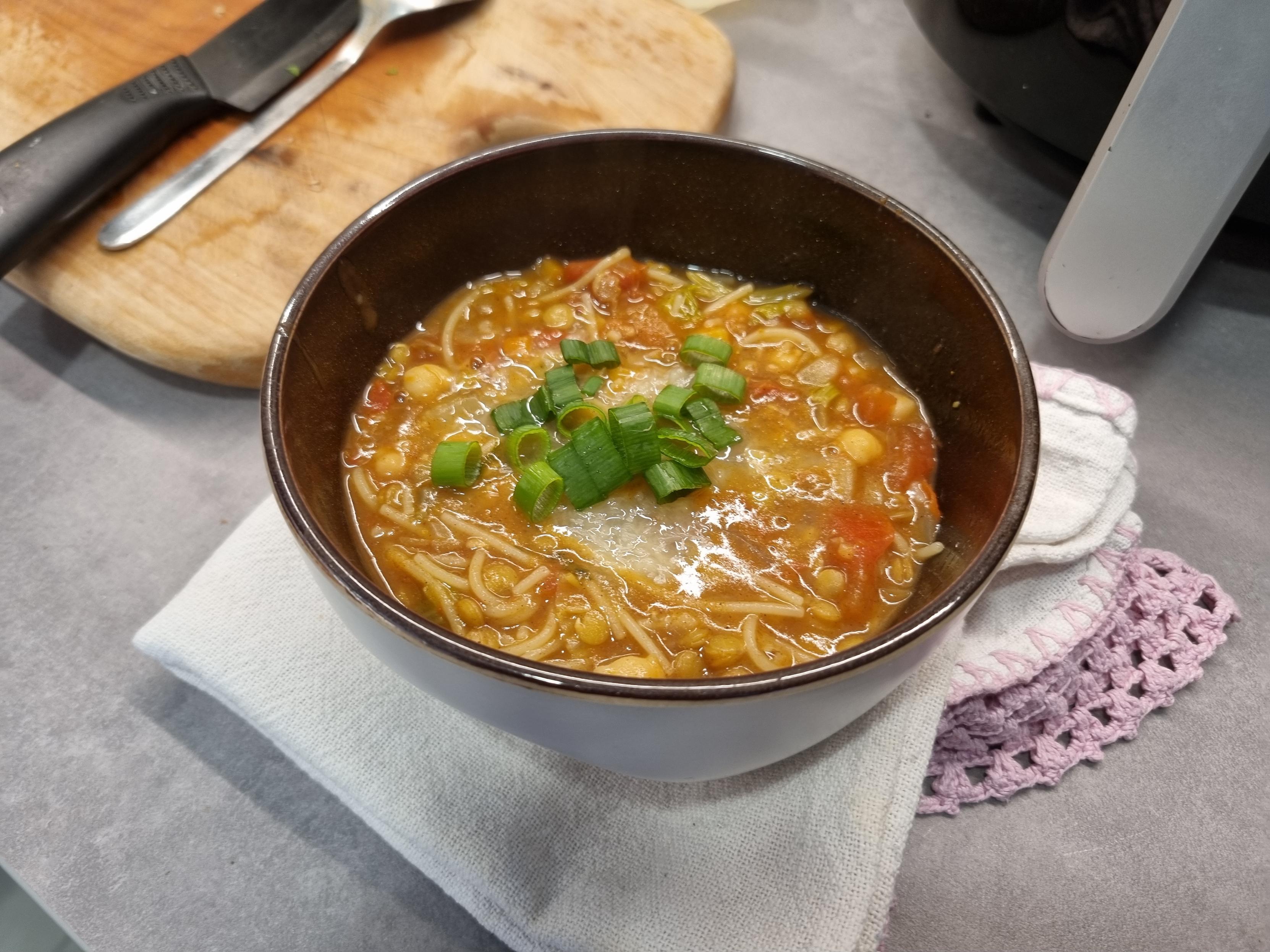 Cumbuca clara com interior marrom brilhoso, em cima de um pano de prato branco e um balcão de cozinha cinza claro. Contém uma sopa vermelha, com tomates, grão de bico, lentilha e massa (Capellini), e cebolinha picada por cima. Ao fundo uma tábua de cortar de madeira com uma faca preta de cerâmica e uma colher em cima.