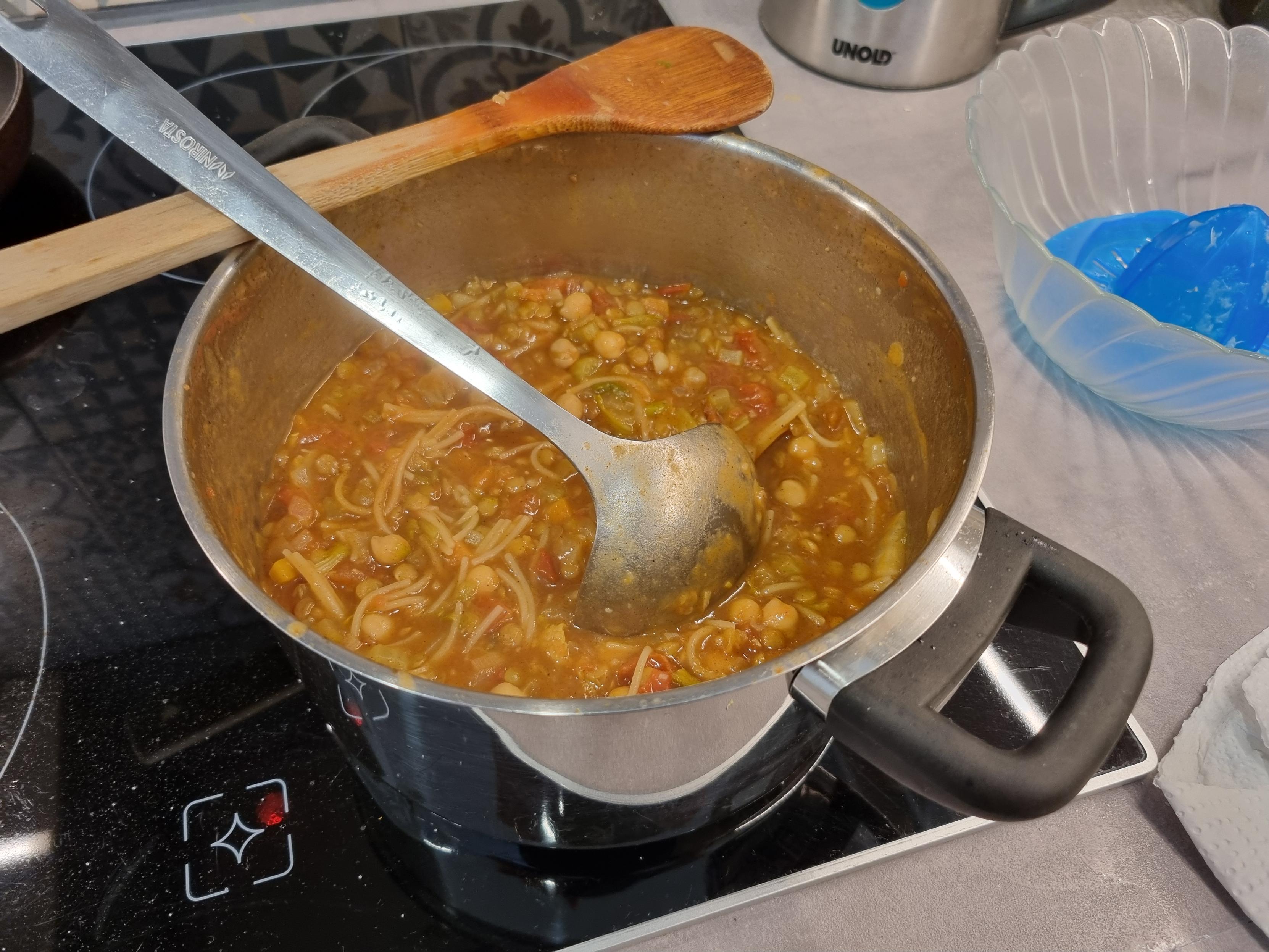 Panela de pressão em cima de um fogão elétrico de cerâmica preta. Contém uma sopa vermelha, com tomates, grão de bico, lentilha e massa (Capellini).
