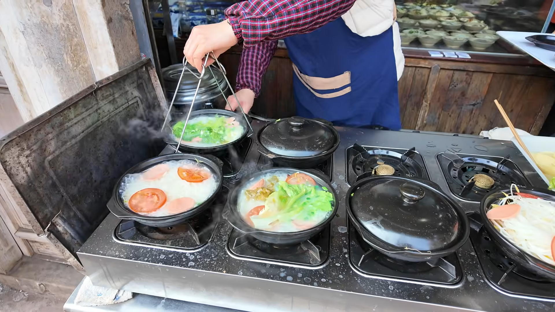 Foto de um frame de vídeo nas ruas da China, mostra várias "panelas" (?) sem cabos com comida sendo preparada; numa delas, uma pessoa usa um pegador metálico para suspendê-la e tirá-la dali.