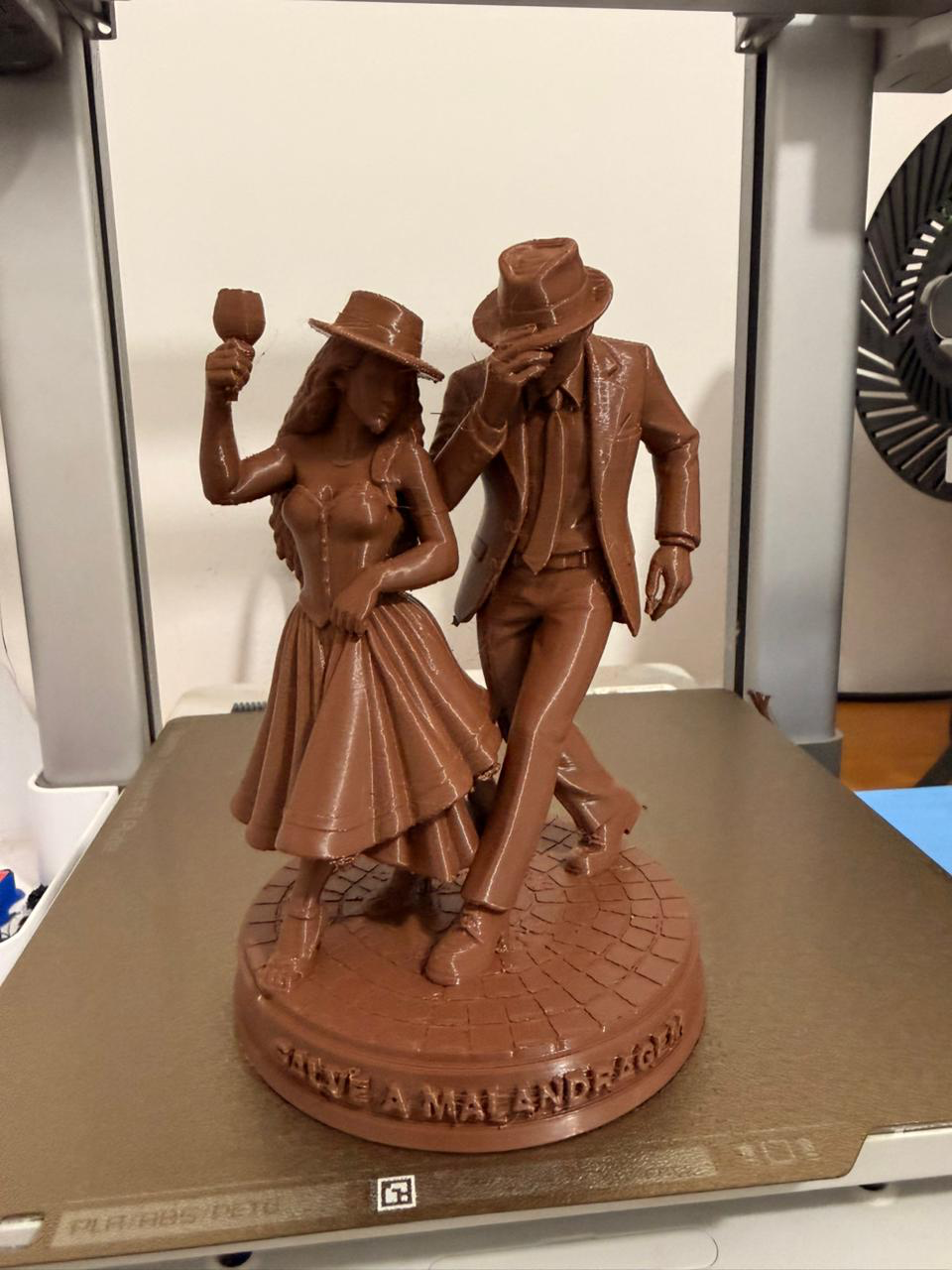 Escultura religiosa de entidades da esquerda da Umbanda, casal de malandros, feita em impressora 3d, escultura está em cima da impressora 3d, a equerda uma mulher com vestido longo segurando uma taça, a direita um hoem de terno e chápeu, dando um passo ao seu lado, os dois estao dançando.