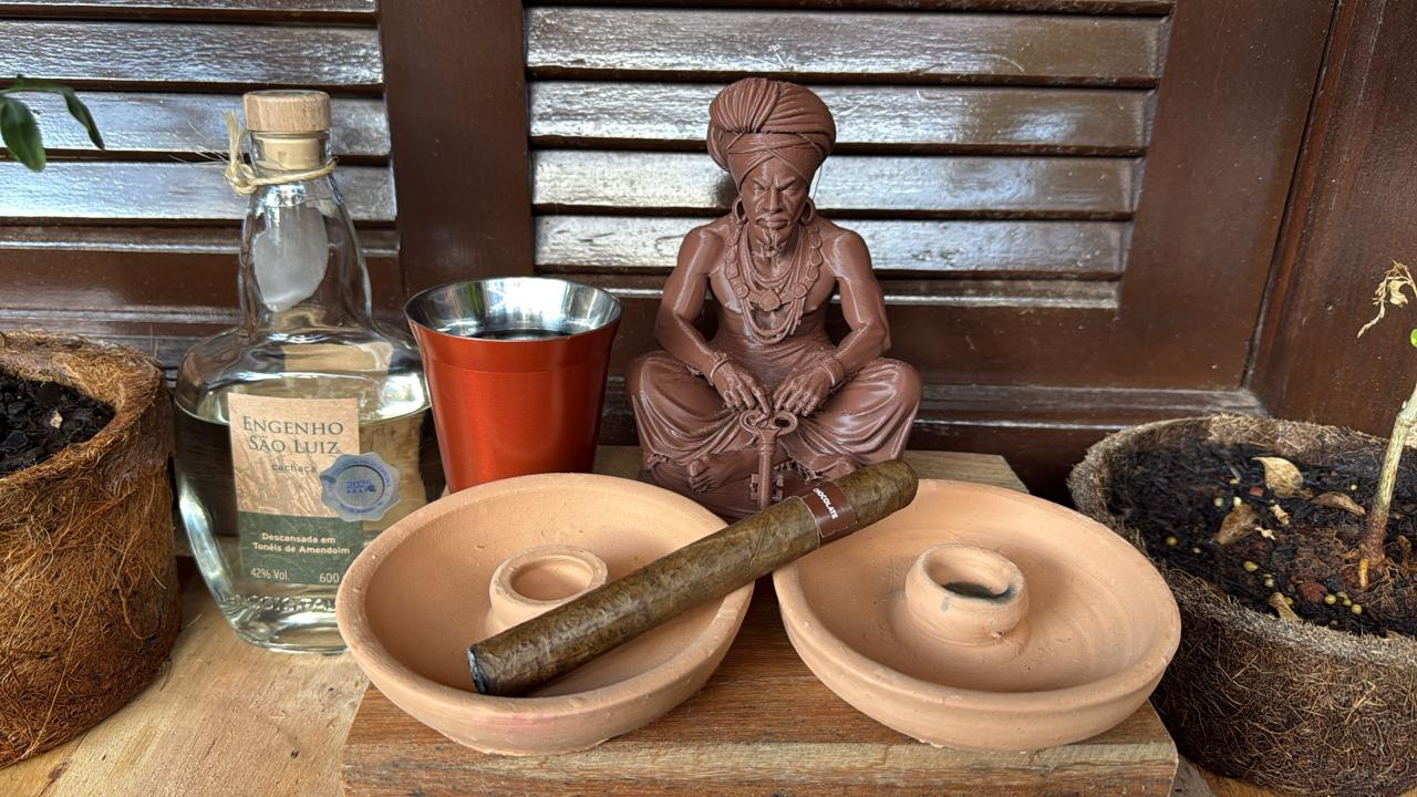 Foto de uma escultura de exu/bará com, um garrafa de marafa do lado esquerdo, um copo com marafa, um charuto cubano e dois suportes de vela a frente, do lado esquerdo uma planta arruda, do direito uma guiné.